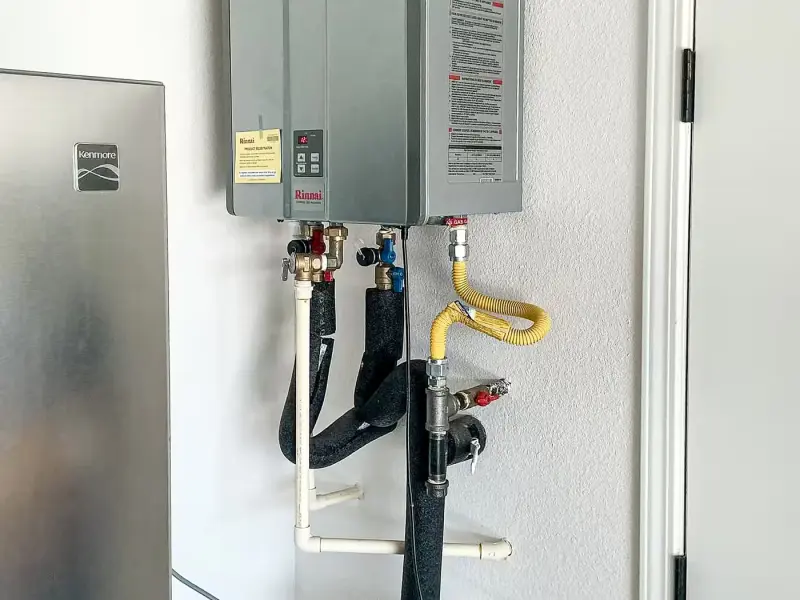 Rinnai tankless water heater on a garage wall with insulated lines and a condensate pump