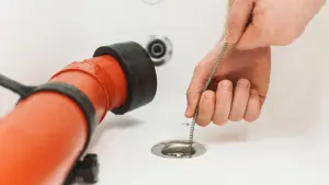 Hand feeding a drain cable into a bathtub drain beside a drain machine hose